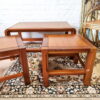 A Mid Century Teak Nest of Tables - Coffee Table and Two Side Tables are arranged on a patterned rug in front of a white brick wall. Two rectangular side tables flank the larger rectangular coffee table. A gold-framed mirror and a small decorative piece are visible in the background.