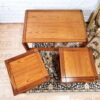 The Mid Century Teak Nest of Tables, consisting of one coffee table and two side tables, are displayed on a light wood floor against a white brick wall. Arranged on top of a small, decorative patterned rug, the rectangular coffee table is complemented by the two square side tables. A part of a framed mirror can be seen to the right.