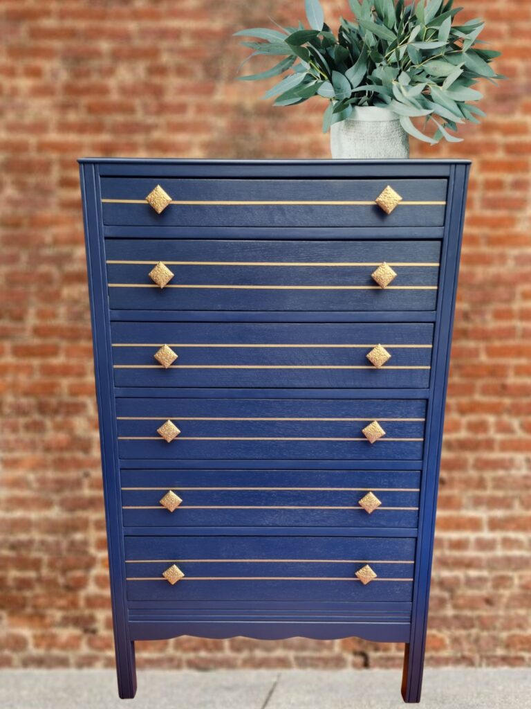 A tall navy blue dresser with seven drawers is adorned with gold diamond-shaped knobs. A green potted plant is positioned on top of the dresser. The background features a red brick wall.