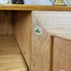 A close-up view of a wooden cabinet corner with a "Made in England" label affixed. The cabinet corner shows detailed craftsmanship with visible wood grain. In the background, part of an interior setting is visible with decorative items.