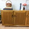 A wooden sideboard with two doors, featuring round handles, stands on a wooden floor against a white brick wall. Decor items, including a basket of flowers, books, and small statues, are placed on top of the sideboard. A circular logo is visible in the upper left corner.