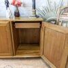 A wooden cabinet with an open door revealing an empty storage space and a small pull-out drawer. On top of the cabinet are decorative items including a lamp, a vase of flowers, and a framed mirror. The setting appears to be indoors with a white brick wall in the background.