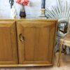 A wooden cabinet with two doors is placed in front of a white brick wall. On top of the cabinet are decorative items including a vase with pink flowers. A mirror with a sunburst frame and a chair with a woven seat are to the side.