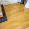 A polished wooden table with a smooth surface. On the left, there is a stack of books and a black object next to them. On the right side, partially visible, is a white textured item, possibly a cloth or napkin. A wall with a light-colored brick pattern is in the background.