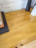 A polished wooden table with a smooth surface. On the left, there is a stack of books and a black object next to them. On the right side, partially visible, is a white textured item, possibly a cloth or napkin. A wall with a light-colored brick pattern is in the background.