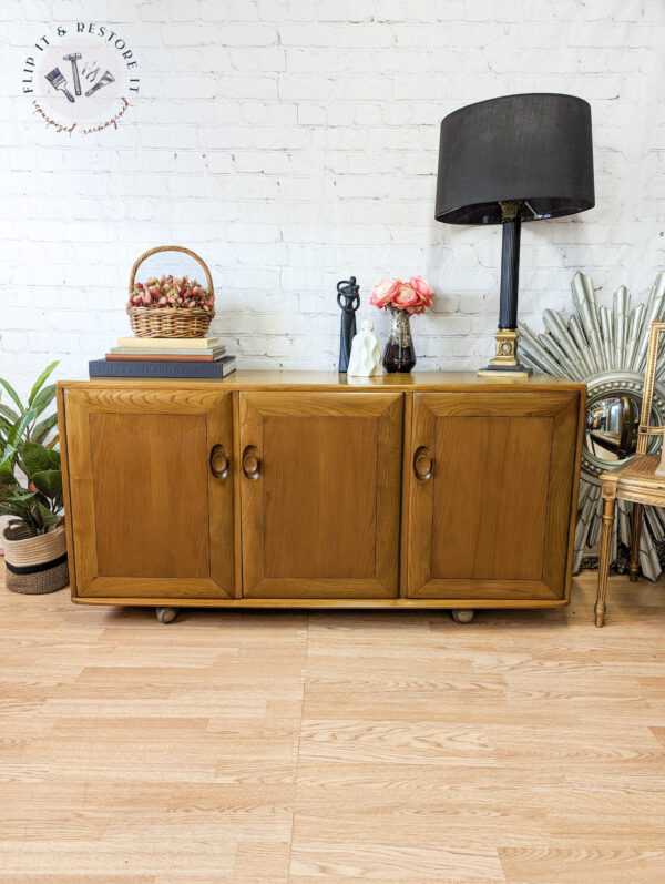 A wooden cabinet with three doors sits against a white brick wall. On top, there's a black lamp, flowers in a vase, and a small basket. To the right, there is a sunburst decor piece and a wooden chair with a white cushion. The floor is light wood.