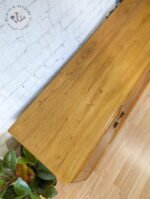 A refinished wooden dresser against a white, textured wall and light wooden floor. The dresser has a smooth, natural finish, showcasing the wood grain. In the bottom left, there's a green plant, adding contrast to the minimalist setup.