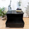 A black wooden bedside table stands against a white brick wall. It has two shelves and an open drawer at the bottom. The top surface displays a giraffe statue, two books, and a small vase with flowers. A potted plant is placed beside the table on the left.