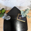 A triangular black wooden table against a white brick wall, topped with a potted plant with silvery leaves, two stacked black hardcover books, and a decorative silver figurine. The table is on a light wood floor and has a "Flip It & Restore It" watermark in the corner.