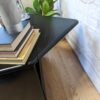 A black triangular table holds a stack of books and a marble vase with greenery. A potted plant is against a light-colored brick wall in the background, and the wooden floor adds warmth to the scene. A circular logo in the top-right corner reads "Flip It & Restore It.