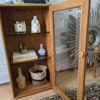 The Ercol Windsor Elm Display Cabinet with a Single Glass Door and Glass Shelves showcases an assortment of items. Inside, there are books, a glass bottle, a white statue, a blue and white vase, and a woven basket. A potted plant sits on top of the cabinet. The cabinet is positioned against a white brick wall beside a decorative mirror.