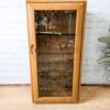 An Ercol Windsor Elm Display Cabinet with a single glass door and two glass shelves stands against a white brick wall. The cabinet is positioned on a wooden floor with part of a patterned rug visible in the foreground. The reflection of someone taking the photo is seen in the glass door.