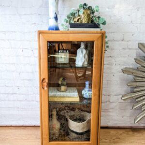 An Ercol Windsor Elm Display Cabinet with a single glass door and glass shelves stands against a white brick wall. Inside are various decorative items, including books, small vases, and a basket. A blue and white vase with flowers sits on top of the cabinet. A fringed rug is partially visible on the floor.