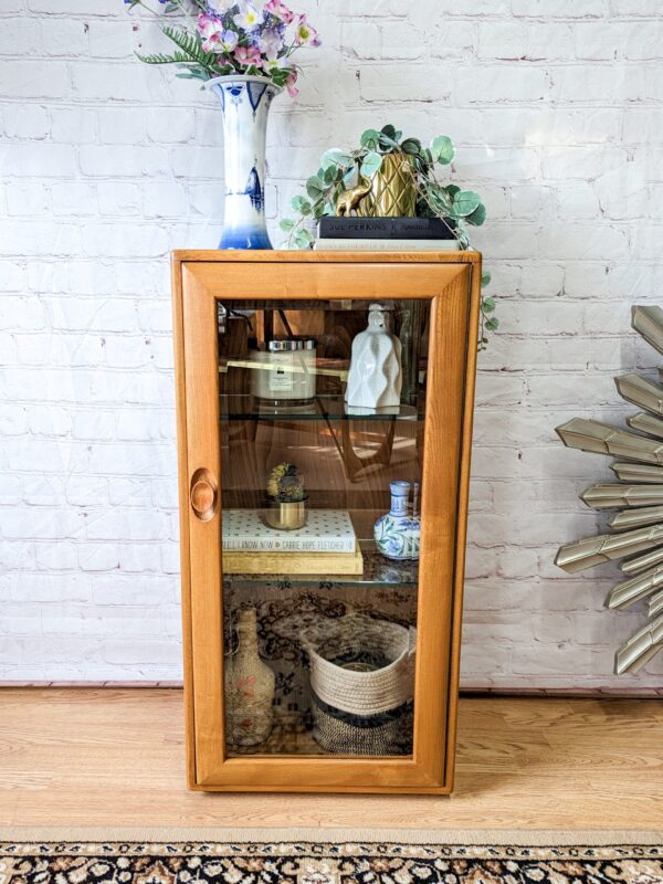 An Ercol Windsor Elm Display Cabinet with a single glass door and glass shelves stands against a white brick wall. Inside are various decorative items, including books, small vases, and a basket. A blue and white vase with flowers sits on top of the cabinet. A fringed rug is partially visible on the floor.