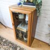 An Ercol Windsor Elm Display Cabinet with a single glass door and glass shelves showcases decorative items, including a patterned book, a small plant, and a woven basket. The top of this wooden cabinet features a blue decorative vase, books, and a potted plant. The cabinet stands on a beige patterned rug.