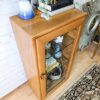 The Ercol Windsor Elm Display Cabinet with a single glass door and glass shelves displays various decorative items. It stands against a light brick wall beside a mirror with a sunburst frame. The top of the cabinet holds books, a ceramic vase, and a plant. A patterned rug lies on the wooden floor in front of it.