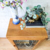 An Ercol Windsor Elm Display Cabinet with a single glass door and glass shelves stands on a patterned rug. On top, there's a tall blue-and-white vase with pink and purple flowers, a small plant with round green leaves in a golden pot, two books, and a small golden figurine of a pig.