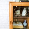 An Ercol Windsor Elm Display Cabinet, featuring a single glass door and glass shelves, showcases various items including books, a white vase, and blue-and-white pottery. Positioned on top of the cabinet are several books and a potted plant. The scene is set against a white brick wall backdrop.