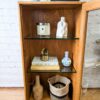 An Ercol Windsor Elm Display Cabinet with a single glass door and glass shelves showcasing various items: a candle, pottery, a stack of books, a small vase, a basket, and a decorative figure. The cabinet is set against a white brick wall and partially open, revealing its contents.