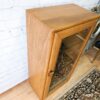 The Ercol Windsor Elm Display Cabinet, featuring a single glass door with reflective shelves and a round knob, stands against a white brick wall. The cabinet rests on a wooden floor next to a carpet adorned with intricate patterns. A brass object is partially visible on the right.