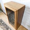 The Ercol Windsor Elm Display Cabinet with a single glass door featuring two interior glass shelves stands against a white brick wall. It has a single knob on the left side of the door and is placed on a wooden floor, partially overlapping an ornate, patterned rug.