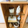 An Ercol Windsor Elm Display Cabinet with a single glass door and two glass shelves. The top shelf holds a candle, a white ceramic figure, and a book. The bottom shelf displays a stack of books, a decorative jar, and a small plant basket. The cabinet has additional books and potted plants on top.