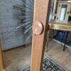 Close-up of the side hinge and latch mechanism of an Ercol Windsor Elm Display Cabinet Single Glass Door with Glass Shelves. The cabinet stands on a wooden floor, and a patterned carpet is partially visible below. In the background, a golden framed mirror and part of a lamp are also visible.