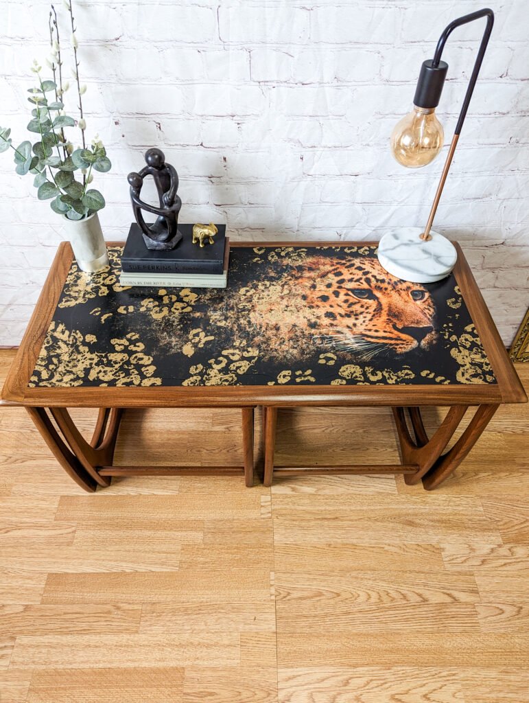 A wooden table with a black surface featuring a leopard's face and golden floral patterns. On top, there's a decorative sculpture, a small potted plant, and a lamp with a marble base and an exposed bulb. The backdrop is a white brick wall, and the flooring is light wood.