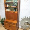 The G-Plan Sierra Tall Teak Bookcase/Drinks Cabinet Mid Century showcases a variety of items with its two lower cabinets and multiple shelves, displaying books, a small statue, a decorative candle, and potted plants. Set against a white brick wall background, the scene is enhanced by the presence of a sunburst mirror on the right.