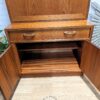 The G-Plan Sierra Tall Teak Bookcase/Drinks Cabinet Mid Century features a single drawer on top and double doors below, which open to reveal an interior shelf. The cabinet boasts a natural wood finish and is situated on a light wooden floor with a logo that reads "Flip it & Restore it.