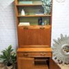 The G-Plan Sierra Tall Teak Bookcase/Drinks Cabinet Mid Century features open upper shelves displaying decor items such as books, a potted plant, a statue, and a "Be Calm" sign. Below is a fold-down desk workspace and a storage compartment with one door ajar. Positioned to the right on the floor is an elegant decorative mirror.