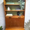 A G-Plan Sierra Tall Teak Bookcase/Drinks Cabinet Mid Century against a white brick wall features open shelves and a closed cabinet at the bottom. The top shelf holds a "Be Calm" sign, book, and potted plant. The middle shelf displays books, a small sculpture, and another plant. Greenery is also visible on the sides of the cabinet.