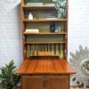 A G-Plan Sierra Tall Teak Bookcase/Drinks Cabinet Mid Century stands against a white brick wall. It features shelves adorned with various decorations, greenery, books, and artistic items. The cabinet door is open, showcasing the stylish interior, while plants decorate the floor to the left and a sunburst mirror is positioned to the right.