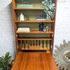 A G-Plan Sierra Tall Teak Bookcase/Drinks Cabinet Mid Century with an open top displaying decorative items such as books, a small vase, a statue, and plants. The bottom section includes vertical slots for organizing papers. The bookcase is surrounded by a decorative mirror, a potted plant, and set against a brick wall background.