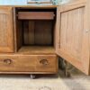 The Ercol Windsor 3 Door 2 Drawer Cabinet Sideboard Model 468 Blue Label Mid-Century features an open wooden design with a drawer at the bottom and an open compartment above it that includes a wooden dowel for hanging items. Placed on a wooden floor, its top is adorned with decorative items like books and plants.