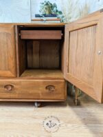 The Ercol Windsor 3 Door 2 Drawer Cabinet Sideboard Model 468 Blue Label Mid-Century features an open wooden design with a drawer at the bottom and an open compartment above it that includes a wooden dowel for hanging items. Placed on a wooden floor, its top is adorned with decorative items like books and plants.