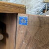 A close-up of the Ercol Windsor 3 Door 2 Drawer Cabinet Sideboard Model 468 Blue Label Mid-Century reveals a small blue and white label in the corner. The label reads "Made in England" with an image of a crown. The wood grain is prominently visible, showcasing its natural patterns and variations.