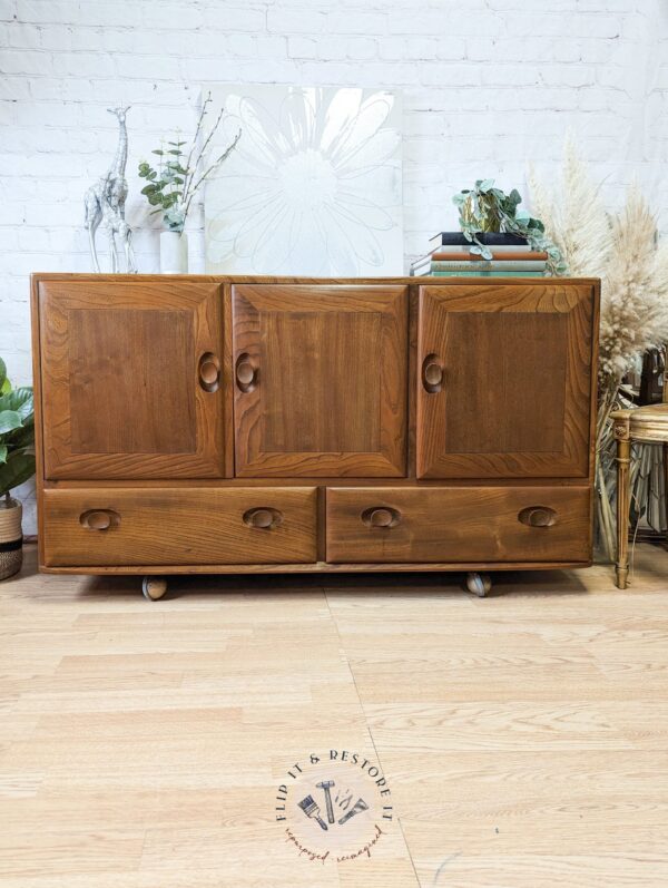 The Ercol Windsor 3 Door 2 Drawer Cabinet Sideboard Model 468 Blue Label Mid-Century sits on a wooden floor against a white brick wall. Decorative items, including a leafy plant, feathered pampas grass, and framed artwork, adorn the sideboard's top.