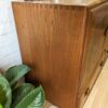 A close-up shot of the Ercol Windsor 3 Door 2 Drawer Cabinet Sideboard Model 468 Blue Label Mid-Century, showcasing its smooth wooden texture and clean lines in a rich brown finish. Decorative items are placed on top, including a metallic object and a plant. Lush green leaves of another plant are visible at the bottom left corner.