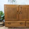 The Ercol Windsor 3 Door 2 Drawer Cabinet Sideboard Model 468 Blue Label Mid-Century stands against a white brick wall, displaying a natural finish. The sideboard features two doors and two drawers, each adorned with oval cutout handles. Decorative items including plants, a giraffe figurine, and sunburst wall art are positioned above it.