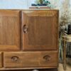 The Ercol Windsor 3 Door 2 Drawer Cabinet Sideboard Model 468 Blue Label Mid-Century stands on a hardwood floor. The sideboard features two large doors with oval handles and a drawer below. A plant and some books are placed on top of the cabinet. Pampas grass and part of a golden chair are visible to the right.