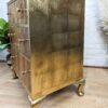 A vintage wooden dresser with a gold-painted finish and four drawers. The dresser stands on four ornate, gold-toned feet. The top and drawer fronts feature a natural wood grain. It's positioned on a hardwood floor with a plant and a white wall in the background.