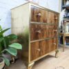 A vintage dresser with a mix of wood and gold finishes. The dresser features three wooden drawers with round brass knobs, ornate gold legs, and decorative plants beside it. A small part of the room's decor and wall art are visible in the background.