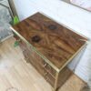 A top-down view of a wooden dresser with a polished, richly grained surface and brass accents. The dresser has three drawers with gold handles and rests on small brass feet. It stands on a light wood floor against a white brick wall, with abstract art nearby.