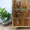 A wooden chest of drawers with gold trim stands on a wooden floor. It has three drawers, each featuring a silver knob. A green leafy plant in a woven basket is placed to the left of the chest. A white brick wall serves as the background.