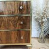 A vintage wooden dresser with three drawers featuring metallic gold trim and round drawer knobs. The dresser stands on carved feet. A pampas grass arrangement in a glass vase is visible to the right on the floor. The background is a white brick wall.