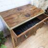 A wooden bar cabinet with an open drawer, revealing wine bottle holders. The cabinet has a polished surface with natural wood grain patterns and brass accents. It sits on a wooden floor next to a white brick wall and decorative plants.