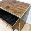 A wooden side table with intricate wood grain patterns, featuring a single open drawer. The drawer's interior is lined with black fabric adorned with a gold scallop pattern. The table is set against a white brick wall and a polished wood floor.