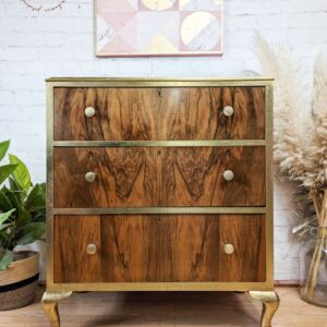 A vintage wooden chest of drawers with three drawers, brass knobs, and gold-painted legs is placed against a white brick wall. Above it hangs a geometric art piece with pastel colors. Plants and dried foliage adorn the surroundings, enhancing the rustic decor.