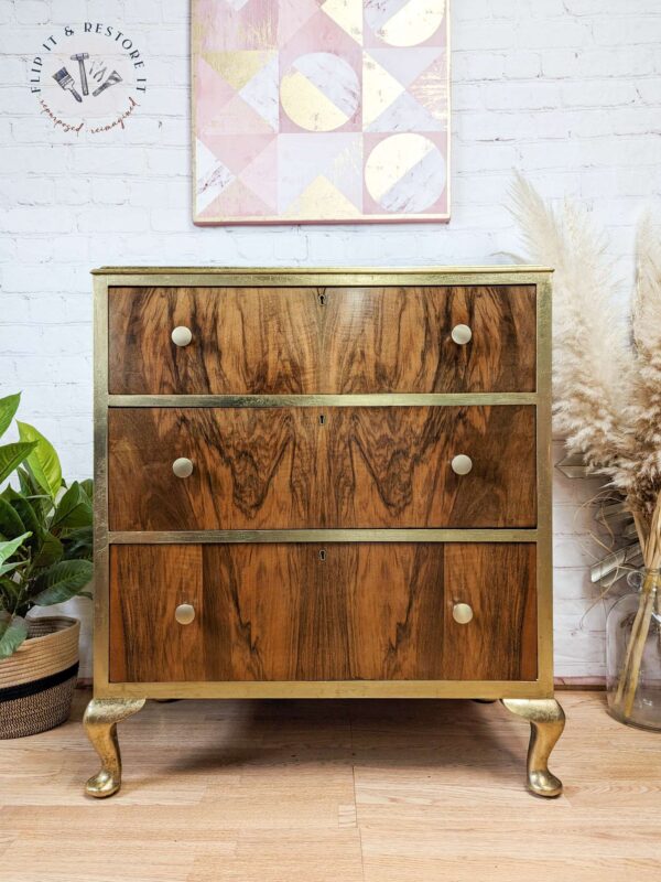 A vintage wooden chest of drawers with three drawers, brass knobs, and gold-painted legs is placed against a white brick wall. Above it hangs a geometric art piece with pastel colors. Plants and dried foliage adorn the surroundings, enhancing the rustic decor.